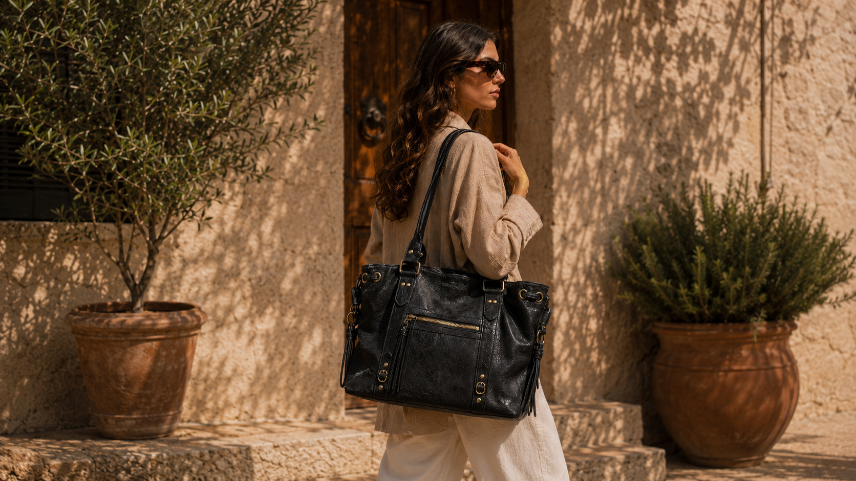 Sac en cuir brossé femme noir à l’aspect patiné porté à l’épaule dans une cour méditerranéenne.